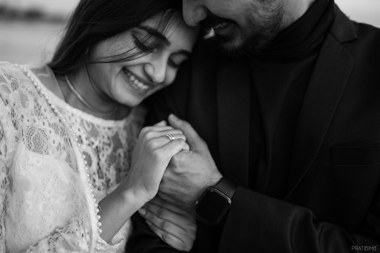 Black and white photo of a smiling couple holding hands, showcasing an engagement ring during a romantic outdoor shoot.