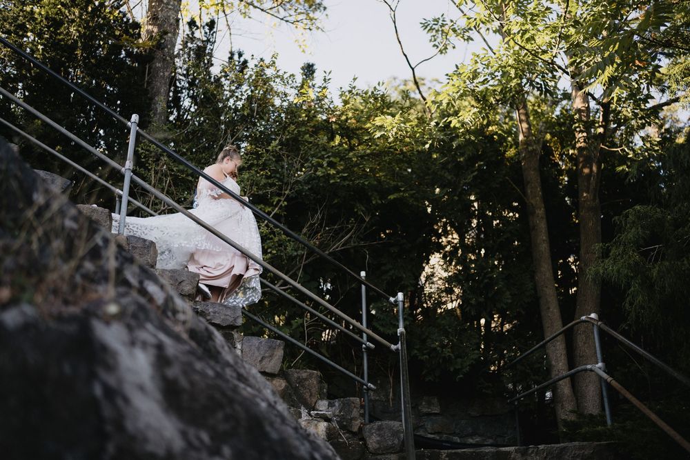 Bride in white gown descending stone steps with metal railing surrounded by lush green trees at outdoor wedding venue.