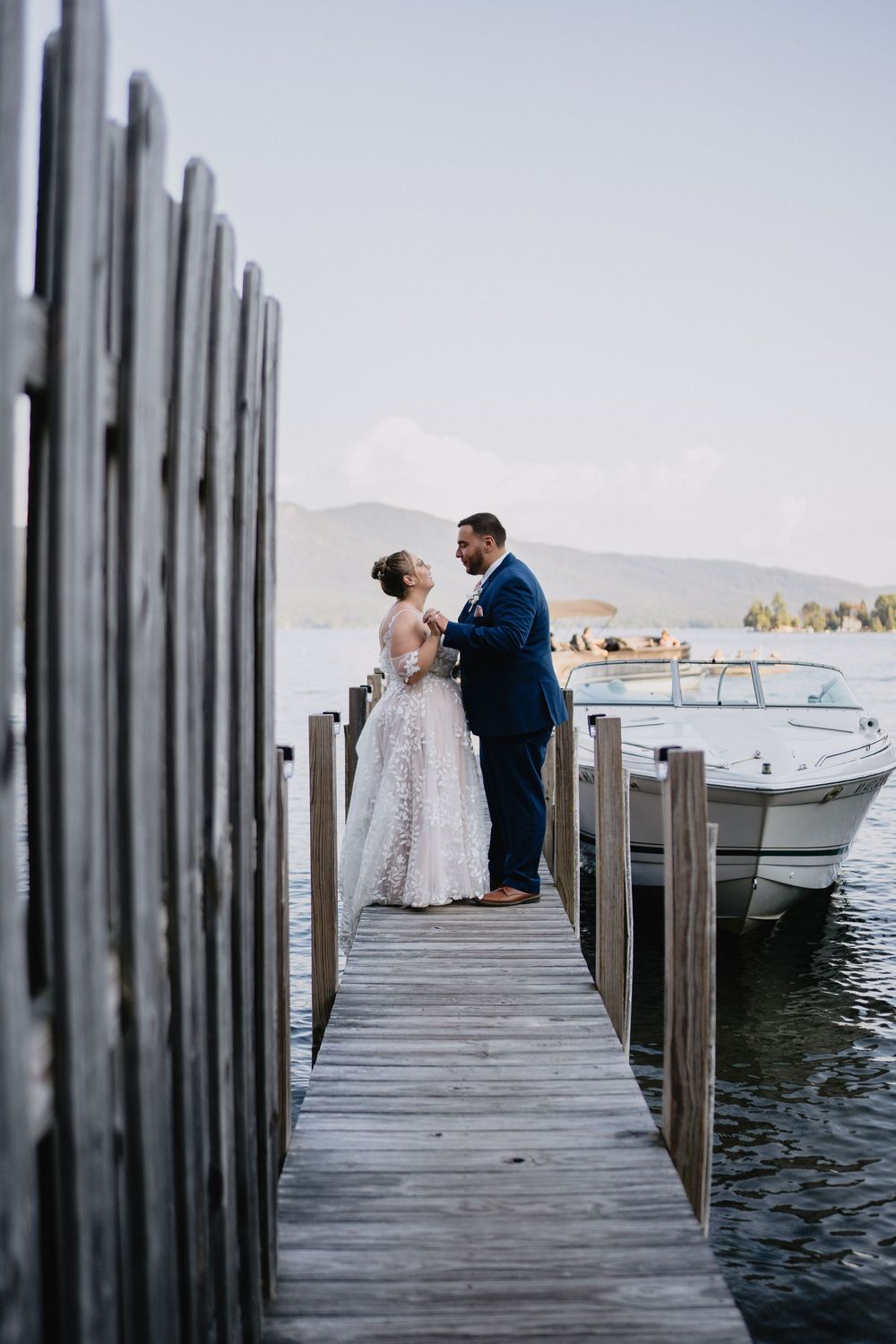 Bride and groom share a romantic moment on a wooden dock by a lake, with a boat and mountains in the background.