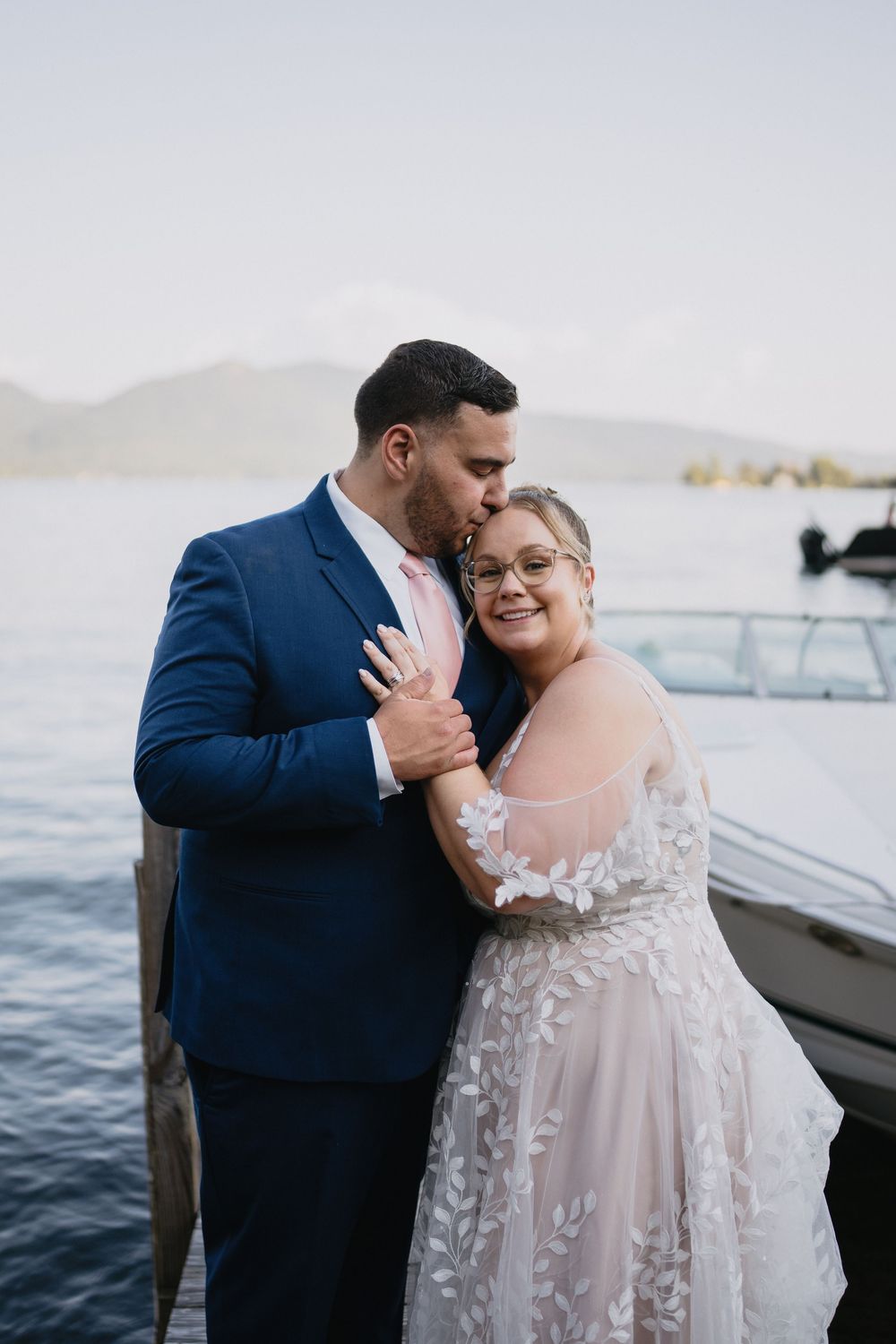 Couple in wedding attire embracing on a lakeside dock, groom in navy suit kissing bride in lace gown.