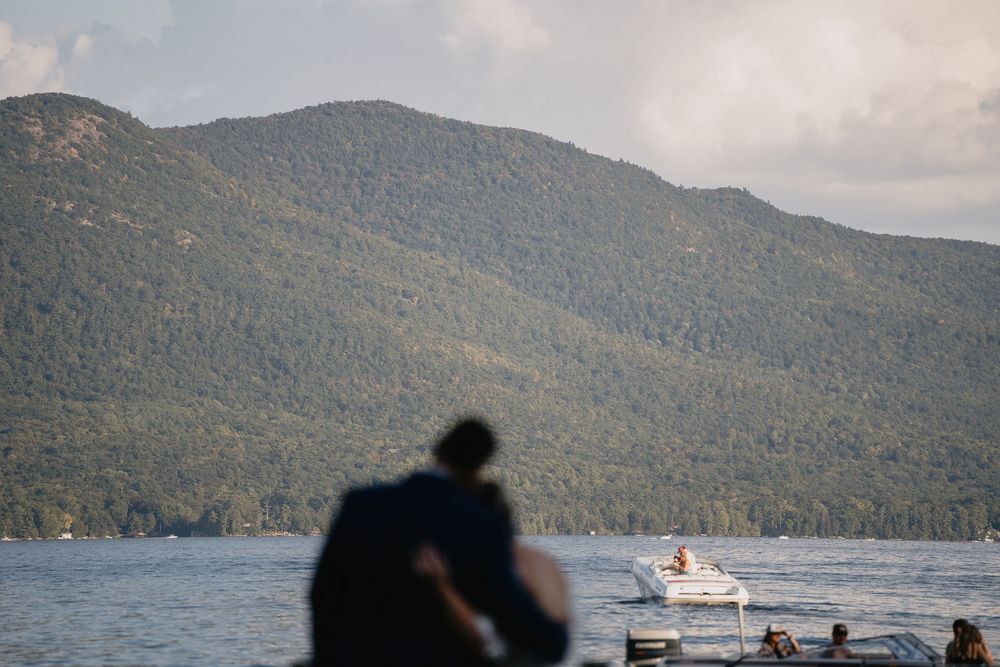 Couple embracing on a lakeside dock with a motorboat and lush green mountain in the background.