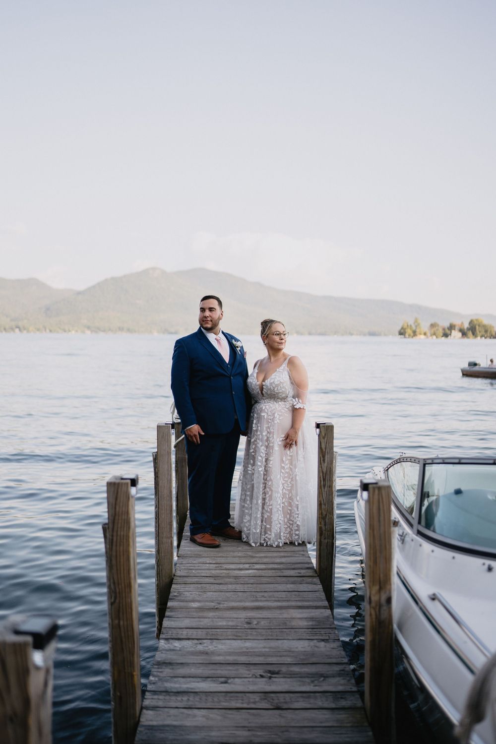 Bride and groom standing on a wooden dock by a lake with mountain views on their wedding day.