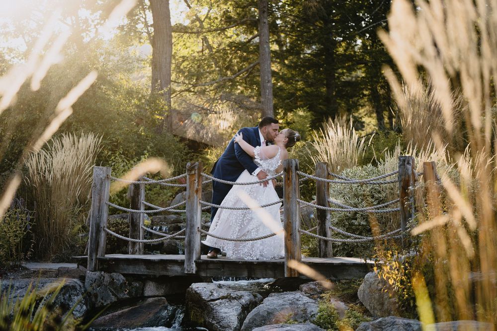 Bride and groom share a romantic dip kiss on a rustic wooden bridge surrounded by lush gardens in golden sunlight.