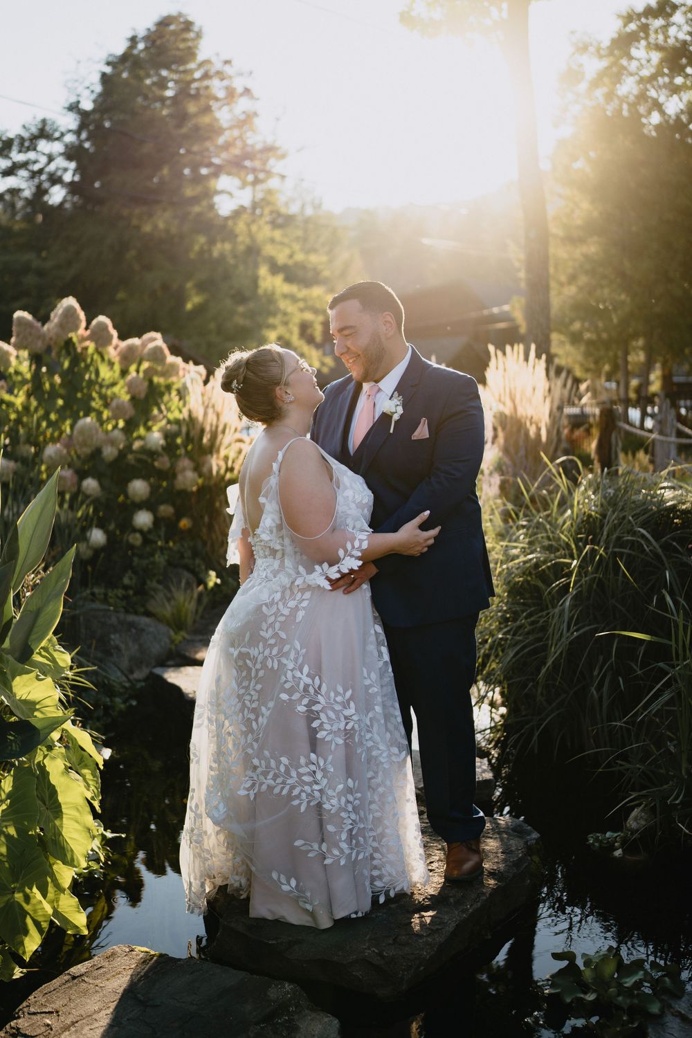 Bride and groom share a romantic moment in a sunlit garden, surrounded by lush greenery and pampas grass near a pond.