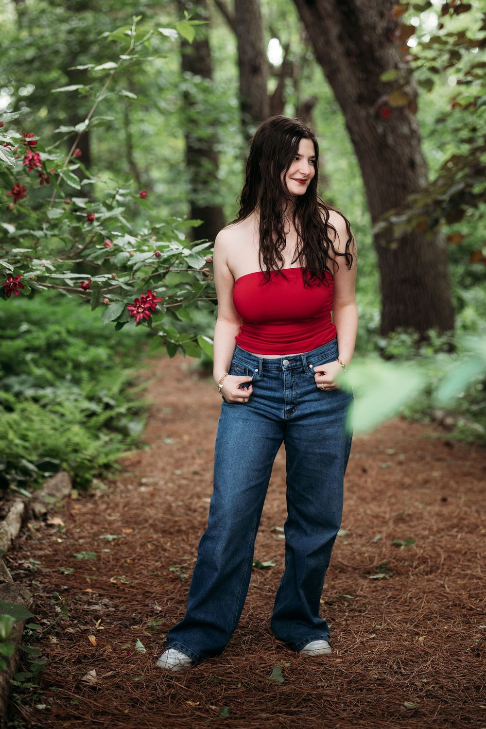 Young woman in red tube top and wide-leg jeans posing on a forest path surrounded by lush green trees.