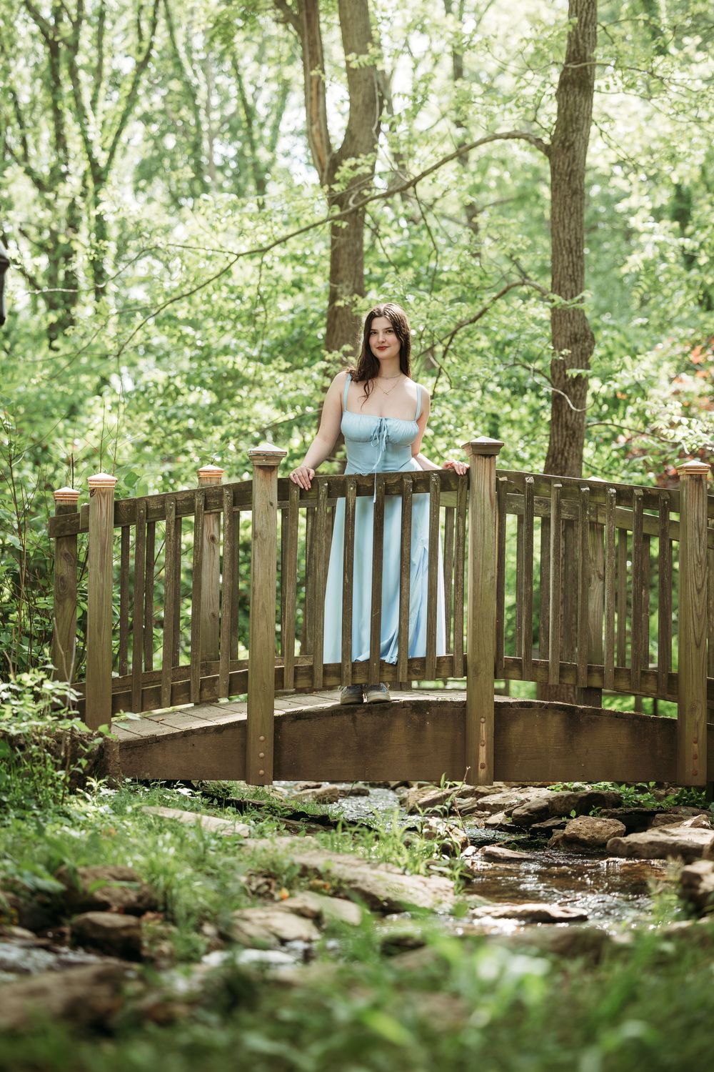A young woman in a light blue dress stands on a wooden bridge surrounded by lush green trees in a serene forest setting.