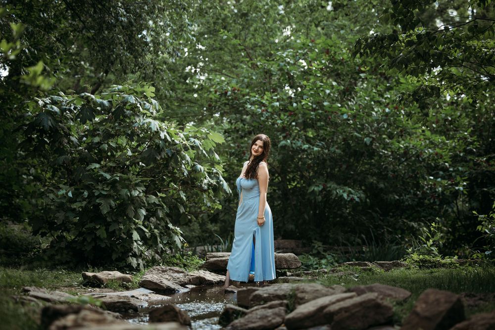 Woman in blue maxi dress standing on rocks surrounded by lush green trees in a serene forest setting.