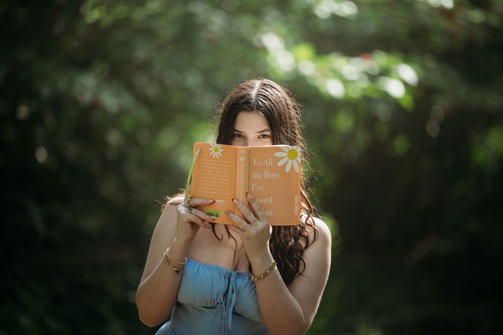 Young woman in blue dress reading a yellow book outdoors, surrounded by lush green bokeh background.