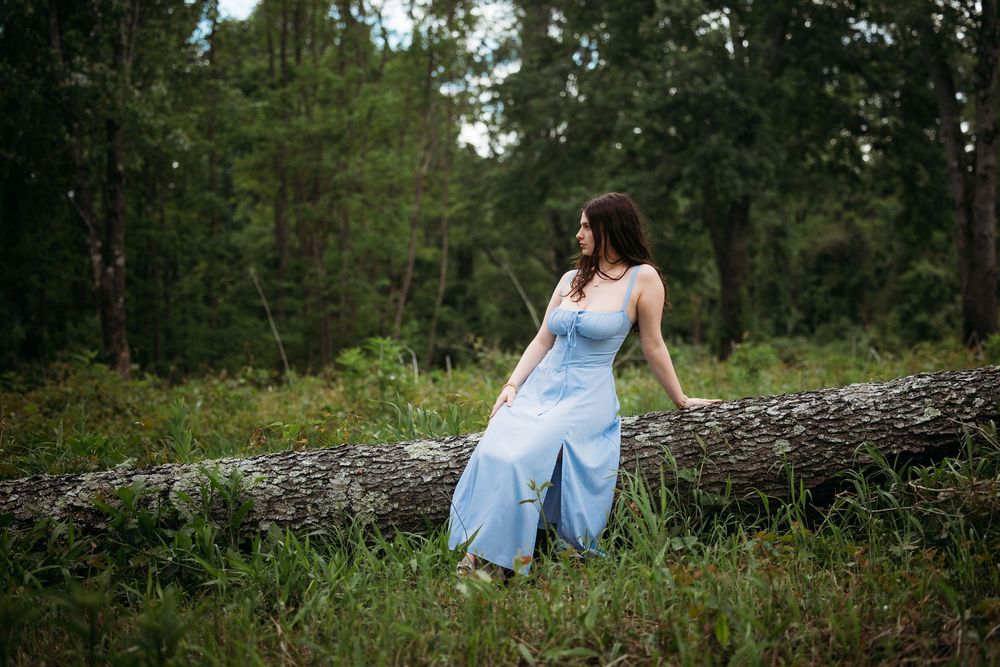 Woman in light blue dress sitting on fallen log in lush green forest setting.