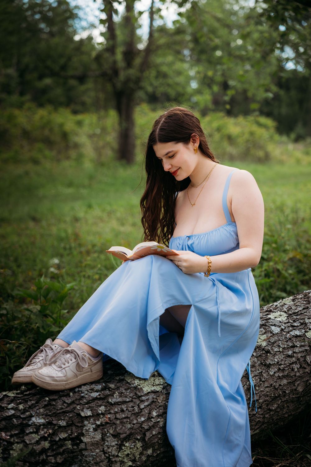 Young woman in blue dress reading a book while sitting on a fallen log in a lush green forest setting.