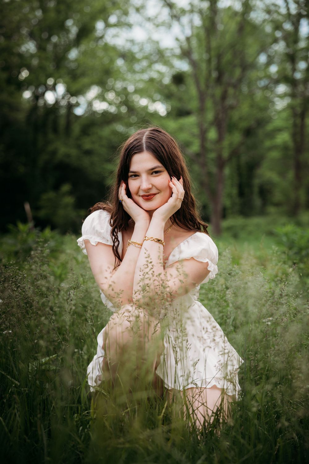 Young woman in white floral dress posing in lush green field, hands framing face, smiling softly at camera.