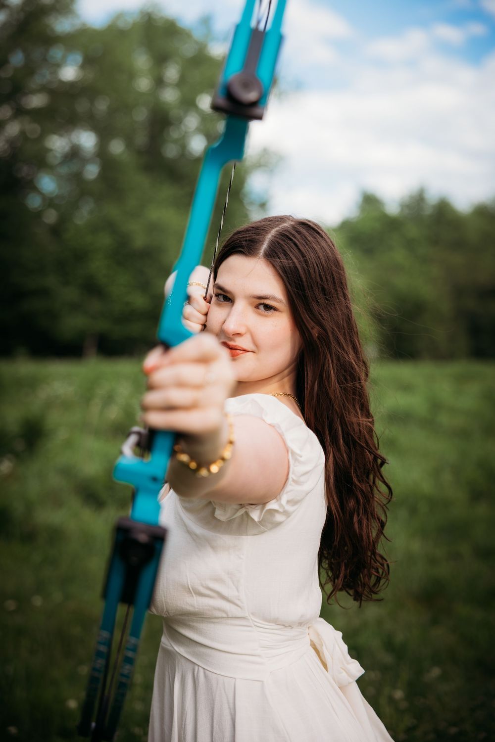 Young woman in white dress aiming a blue recurve bow outdoors in a green field.