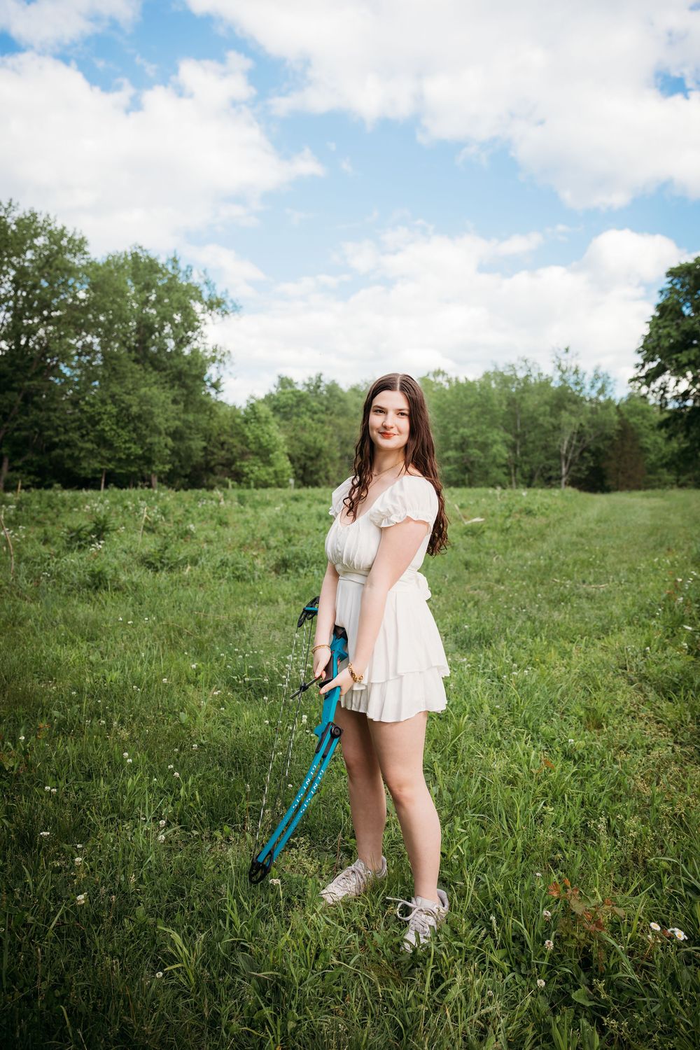 Young woman in white dress holding a badminton racket standing in a green meadow with trees and blue sky.
