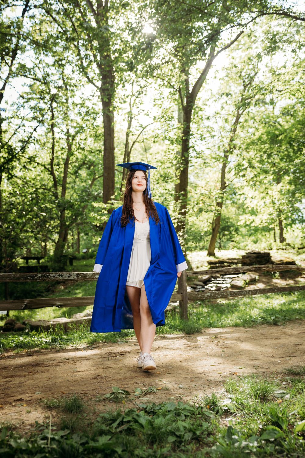 Young woman in blue graduation cap and gown walking outdoors in a lush green forest setting.