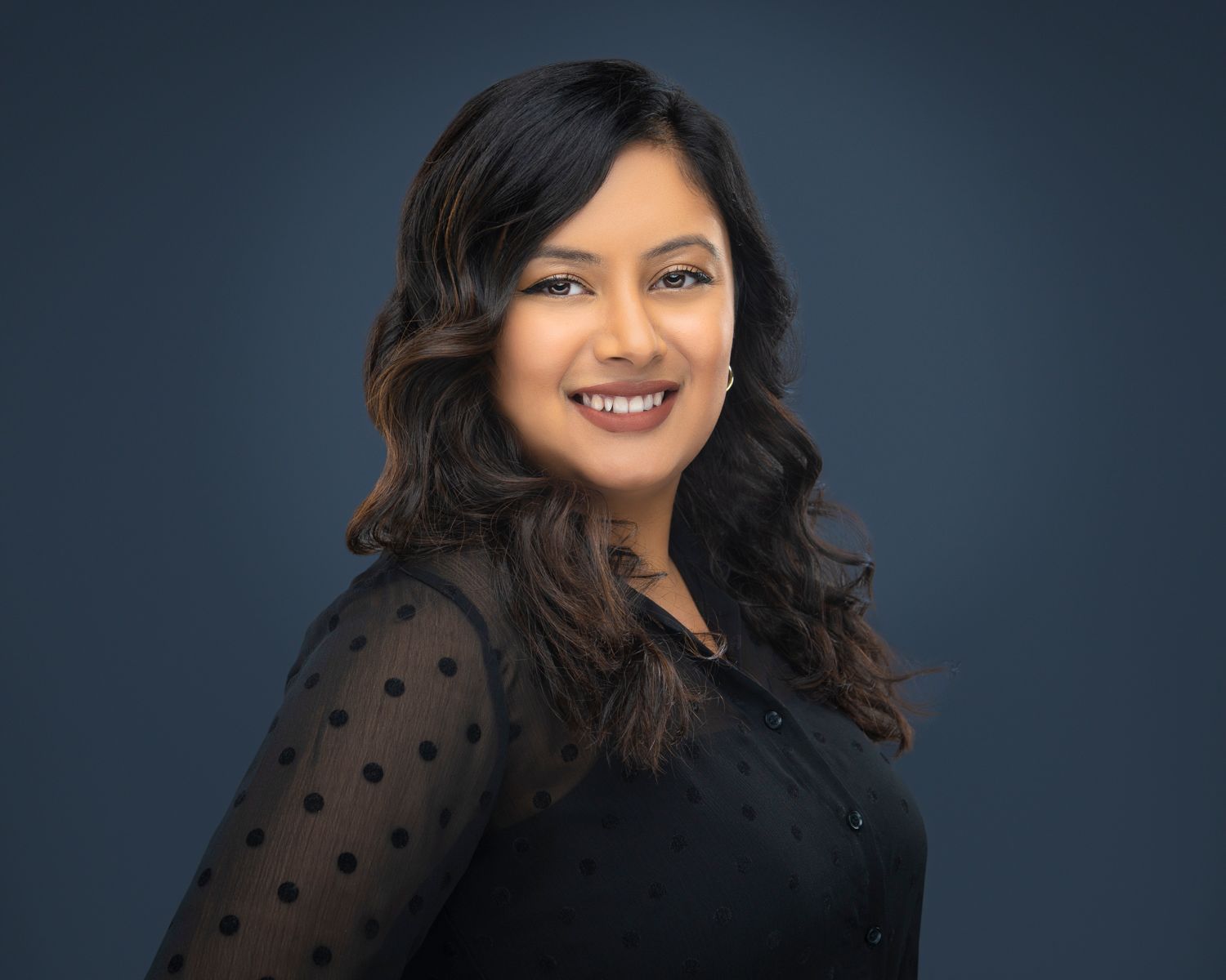Professional headshot of a smiling South Asian woman with wavy hair wearing a black polka dot blouse against a dark blue background.