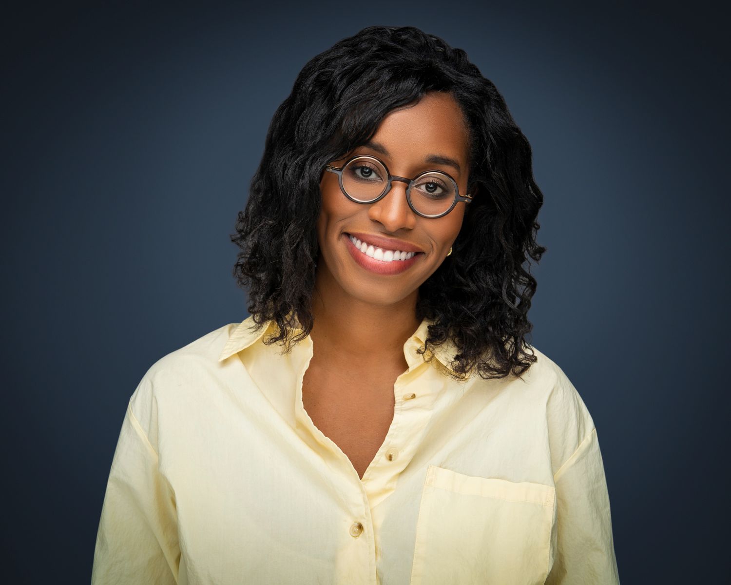 Smiling Black woman with curly hair and round glasses wearing a yellow shirt against a dark blue background.