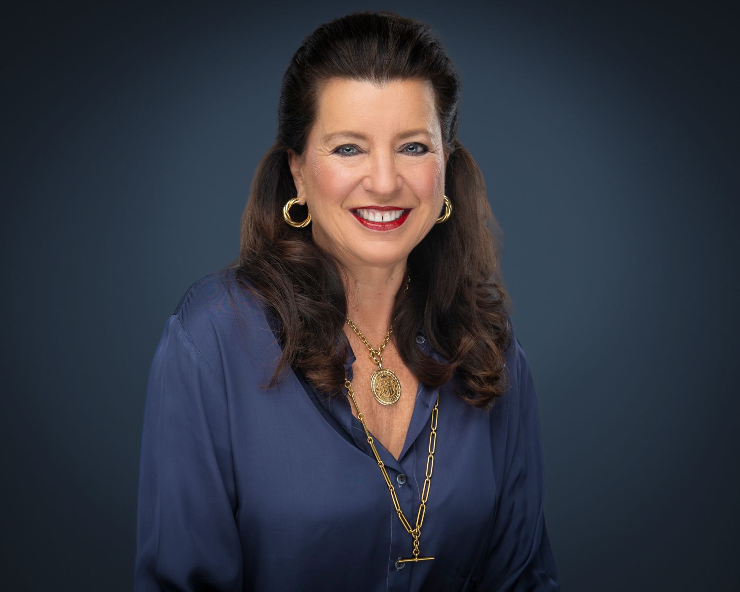 Professional headshot of a smiling woman with dark hair wearing a navy blouse and gold necklace against a dark background.