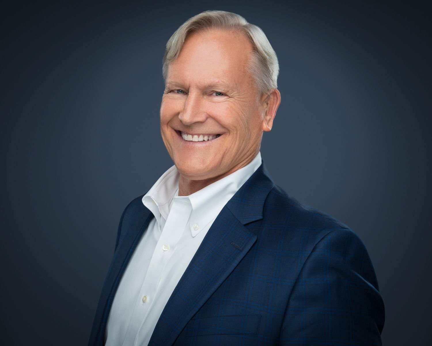 Professional headshot of a smiling middle-aged man in a navy blazer and white shirt against a dark background.
