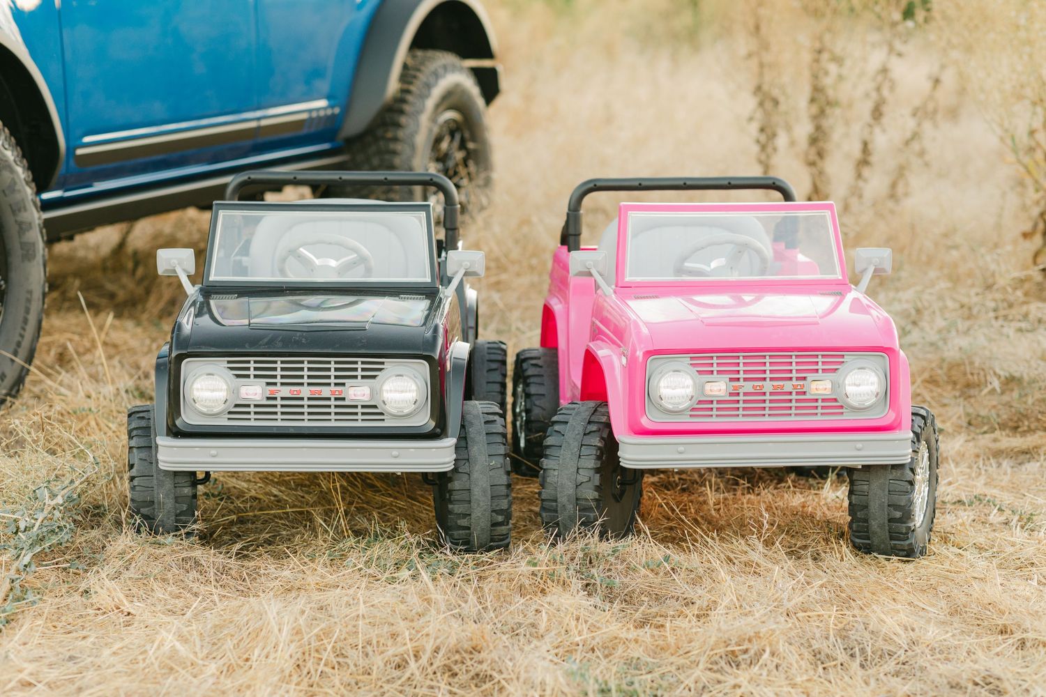 Two toy ride-on SUV trucks, one black and one pink, parked on dry grass next to a blue full-size truck.