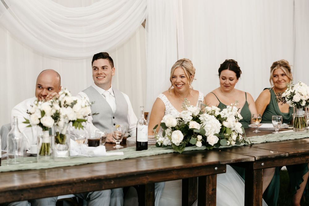 Wedding party smiling at head table with white floral arrangements in elegant white draped reception venue.