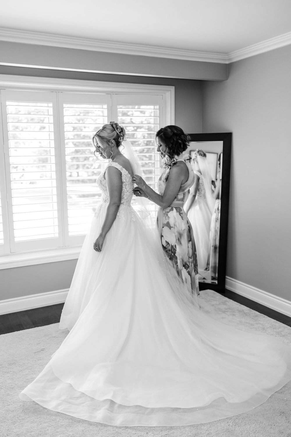 Black and white photo of a bride in a white gown being helped by a woman near a mirror and bright window.