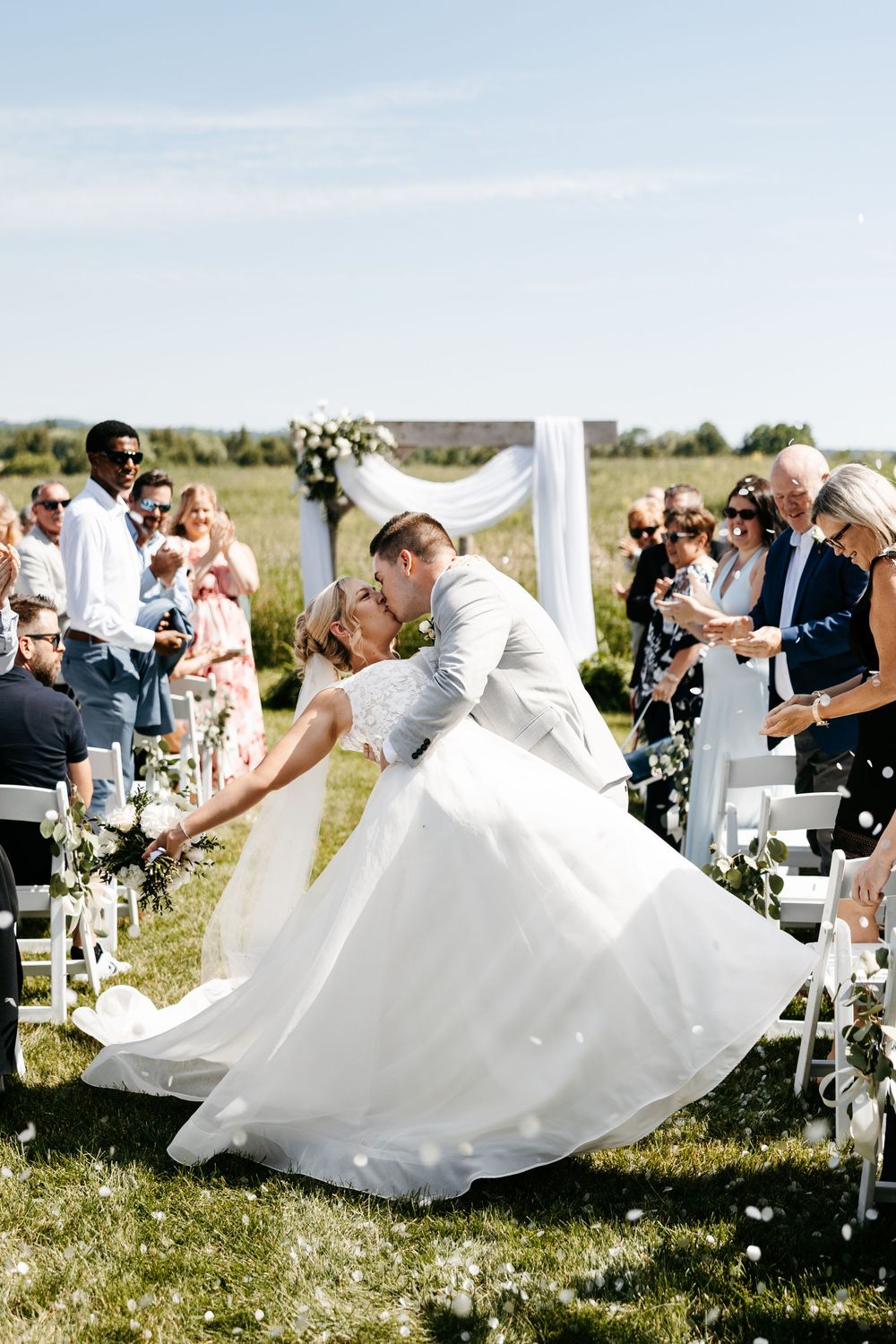 Bride and groom share a romantic dip kiss at outdoor wedding ceremony with white chairs and floral arch.