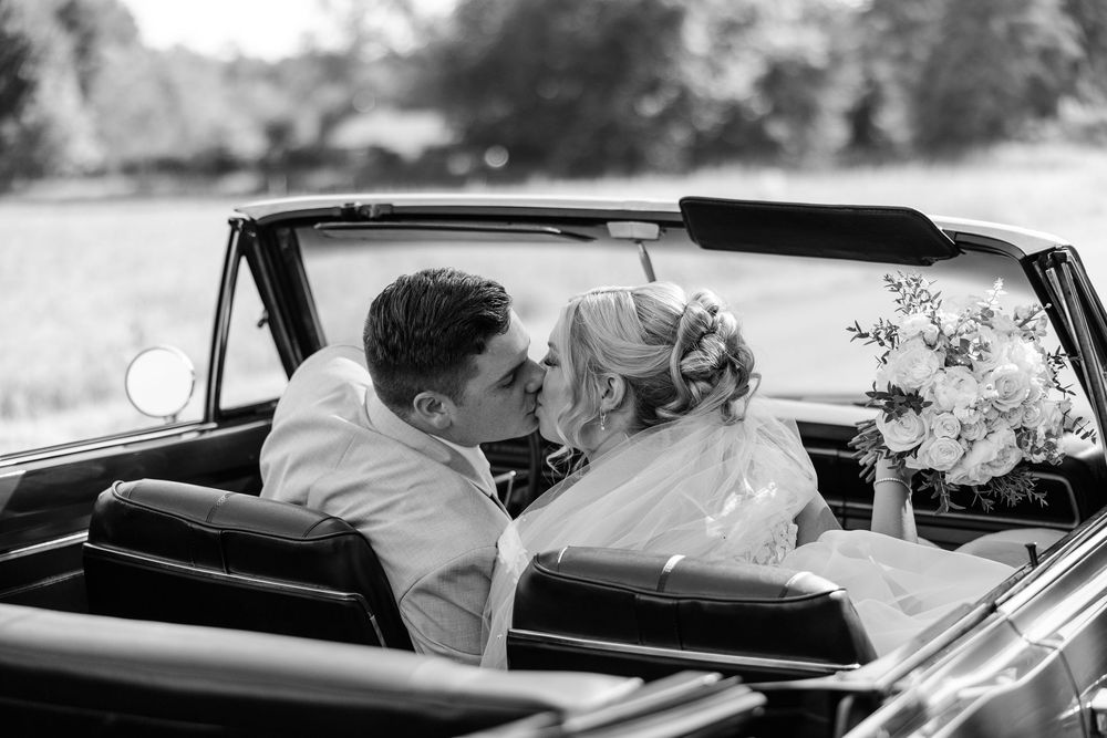 Black and white photo of a bride and groom sharing a kiss in a vintage convertible car on their wedding day.