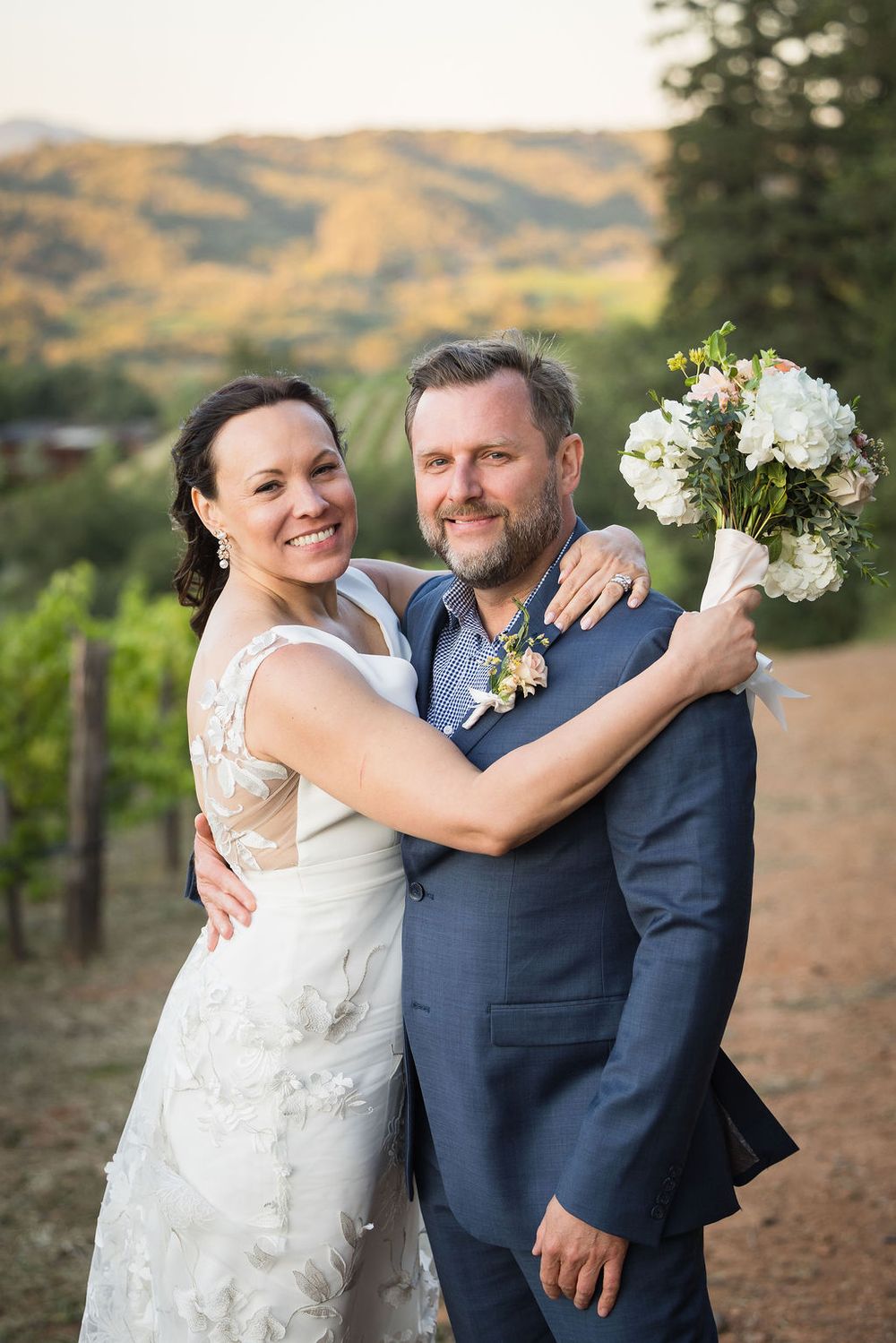 Happy bride and groom pose with bouquet in vineyard with golden hills in background.