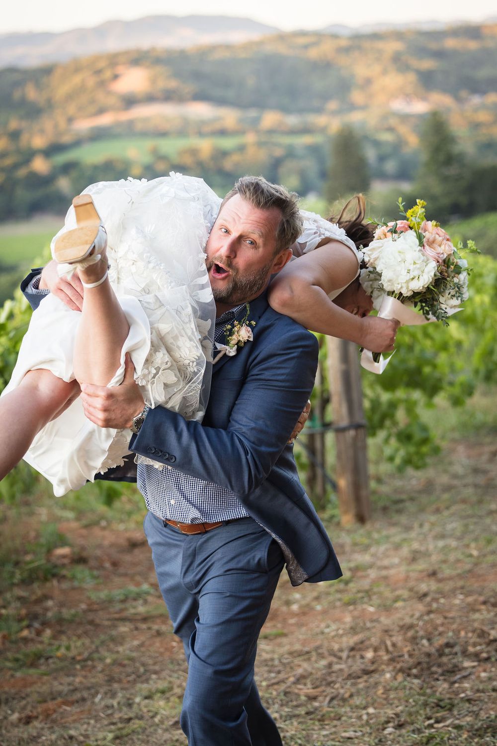 Groom carrying bride over his shoulder in vineyard wedding photo with rolling hills backdrop.