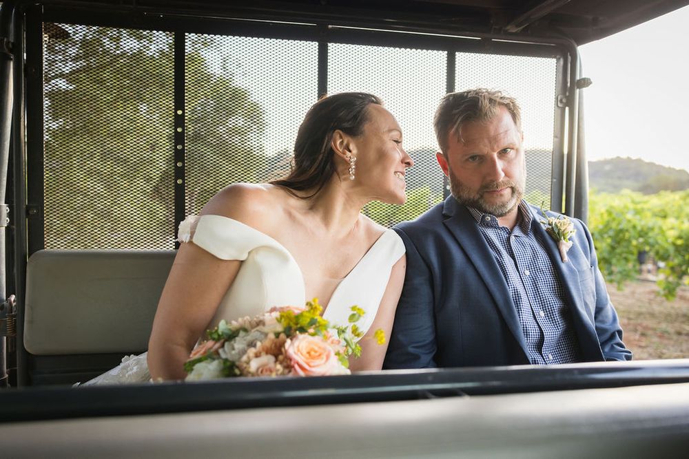 Bride and groom share a joyful moment riding together in a vehicle through a vineyard.