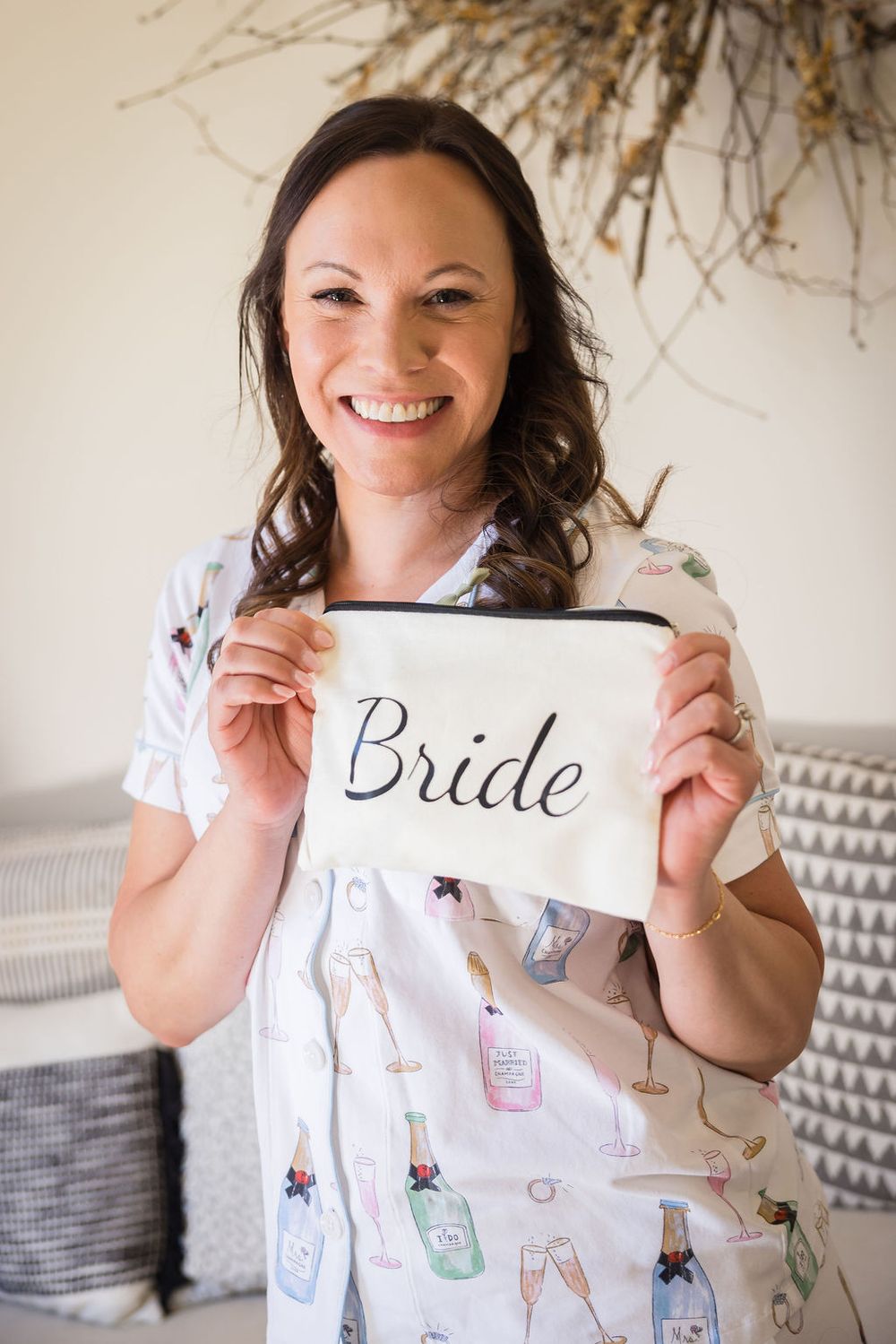Smiling bride holds white cosmetic bag with Bride script text wearing champagne print robe indoors.