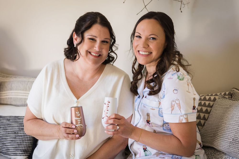 Two smiling women sit together holding beauty products, one in white top and one in floral pajamas, during bridal preparations.