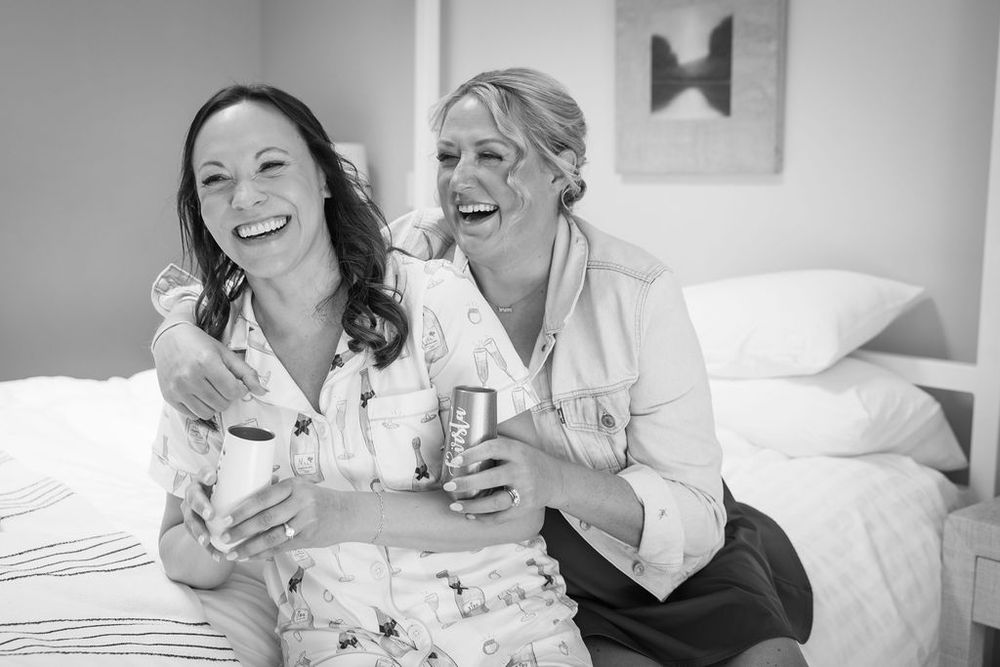 Two women laughing together on a bed holding drinks during wedding day preparations.