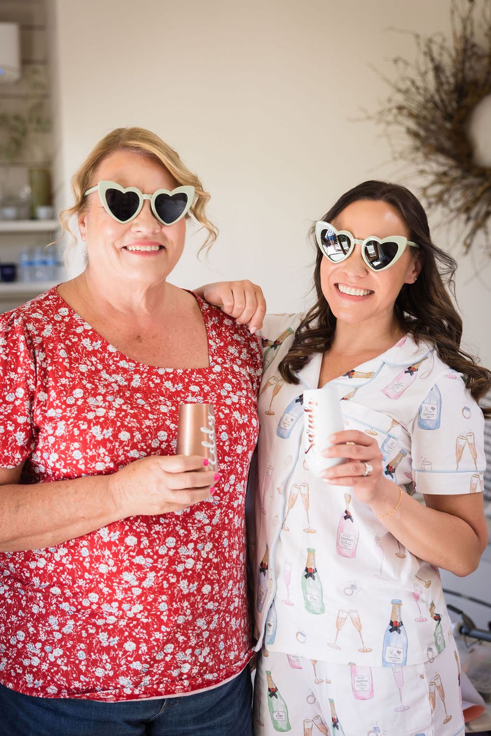 Two women wearing heart-shaped sunglasses smile while holding drinks, dressed in festive attire for a celebration.