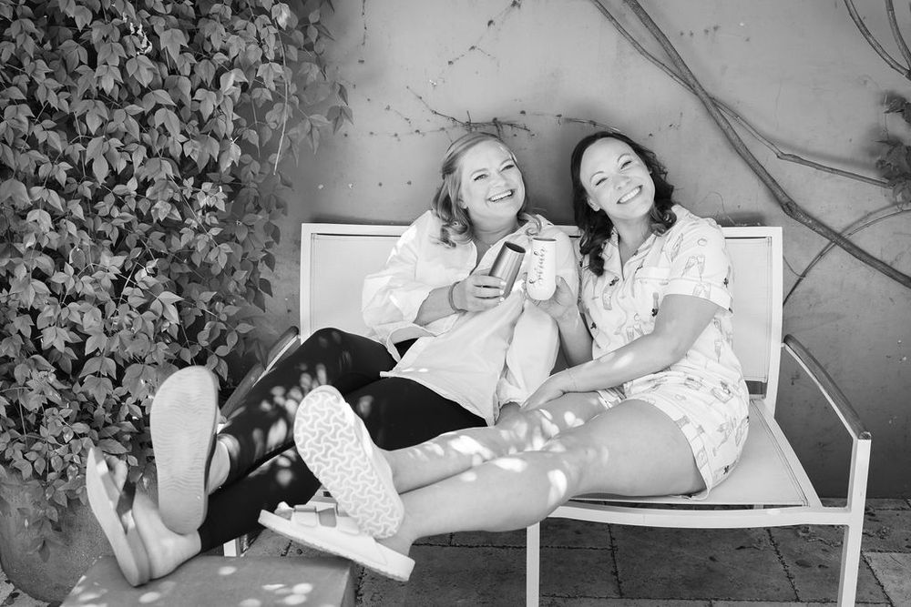 Two women laughing together on white bench beside ivy wall in black and white photo.