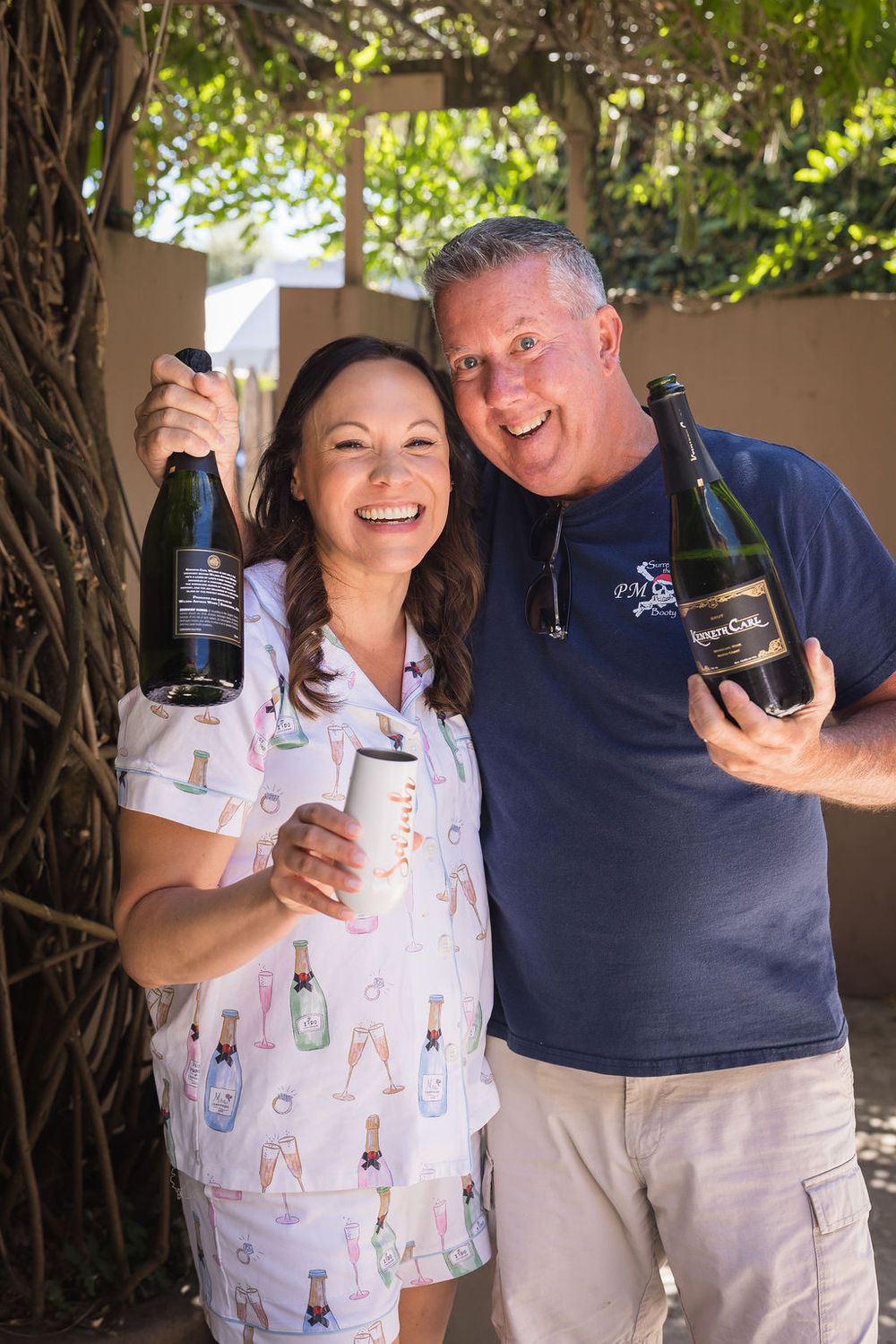 Smiling couple holding wine bottles outdoors, celebrating with champagne near a rustic tree backdrop.