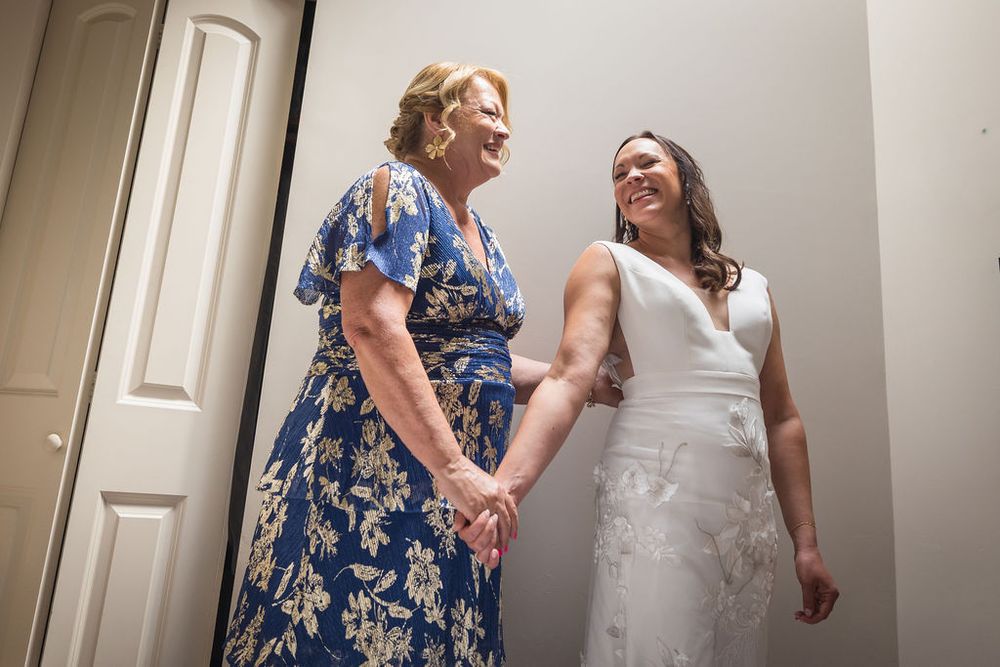 Bride in white lace gown holds hands with older woman in blue floral dress, sharing a joyful moment before the wedding.