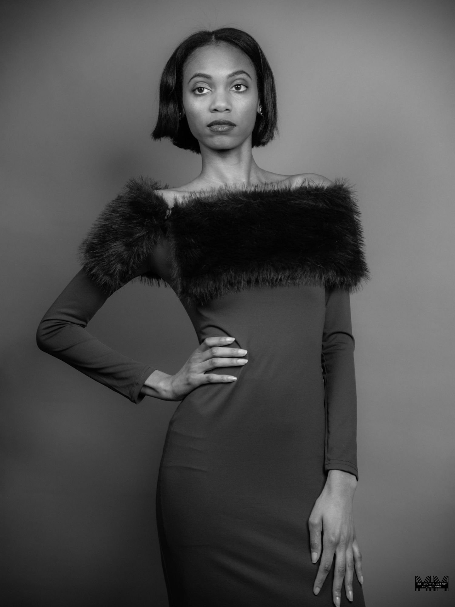 Black and white portrait of a woman in an off-shoulder dress with fur trim, posing confidently in a studio.