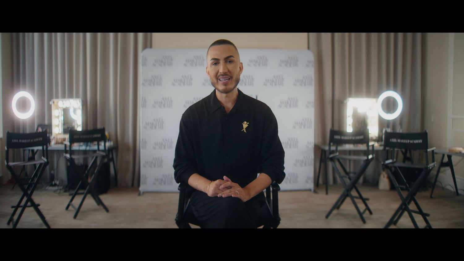 A bald man in a black outfit stands confidently in a studio setting with ring lights and director chairs.
