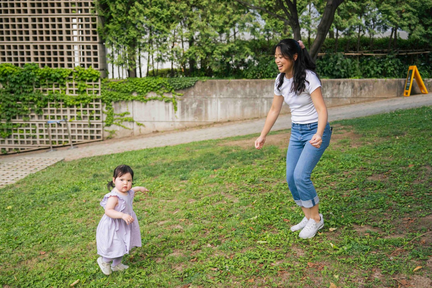 A young mother plays with her toddler daughter in a lush green park, the little girl wearing a pink dress.