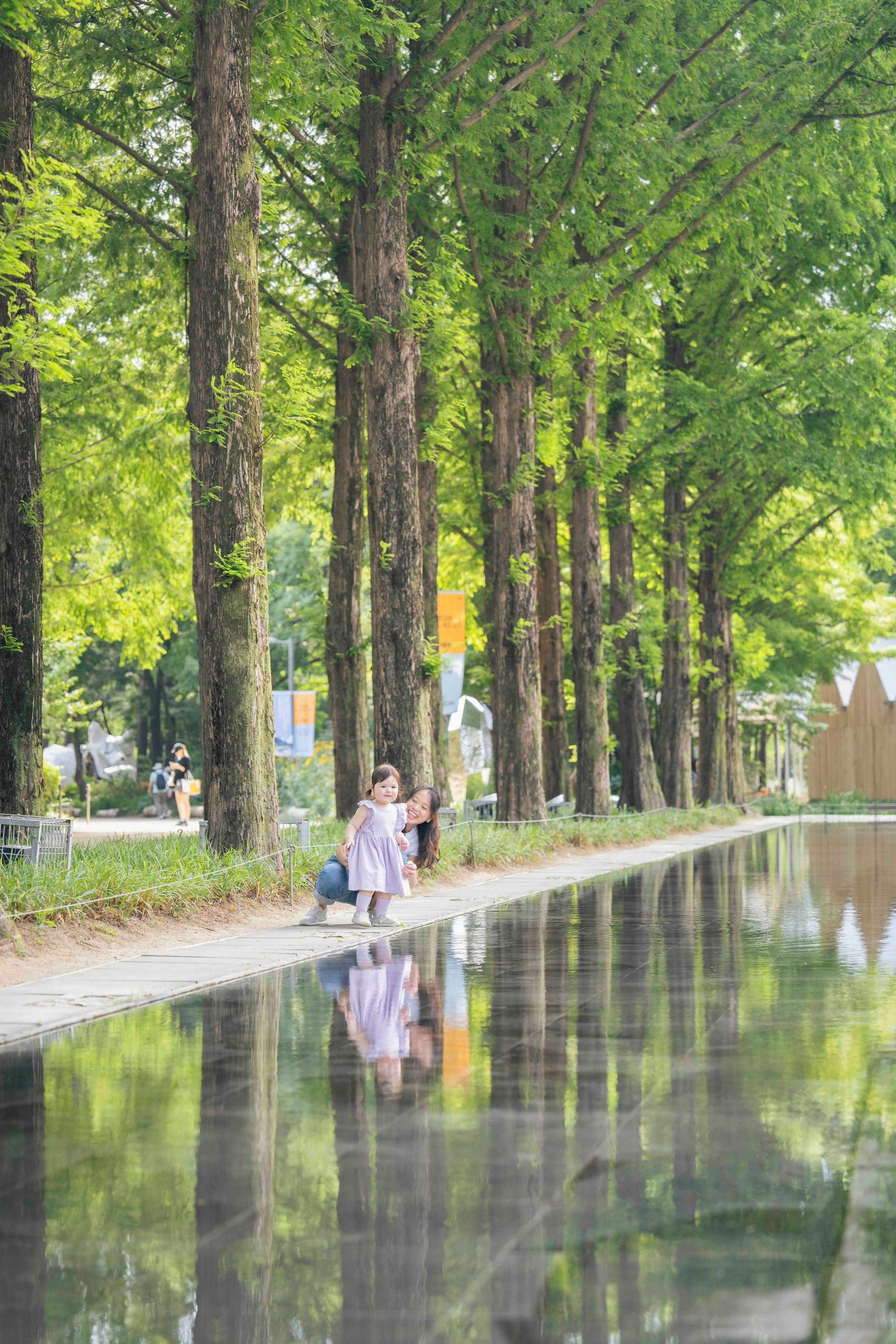 Two women pose along a tree-lined reflective path in a lush green park during summer.