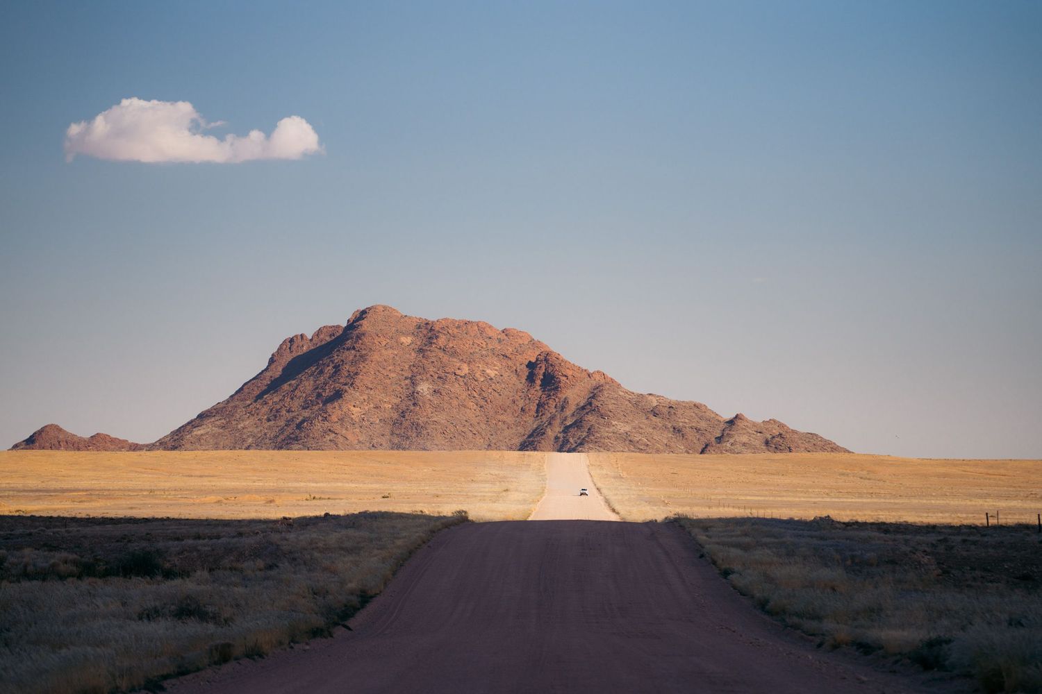 longue ligne droite d'une piste en namibie avec un 4x4 au pied de montagne rocheuse rouge
