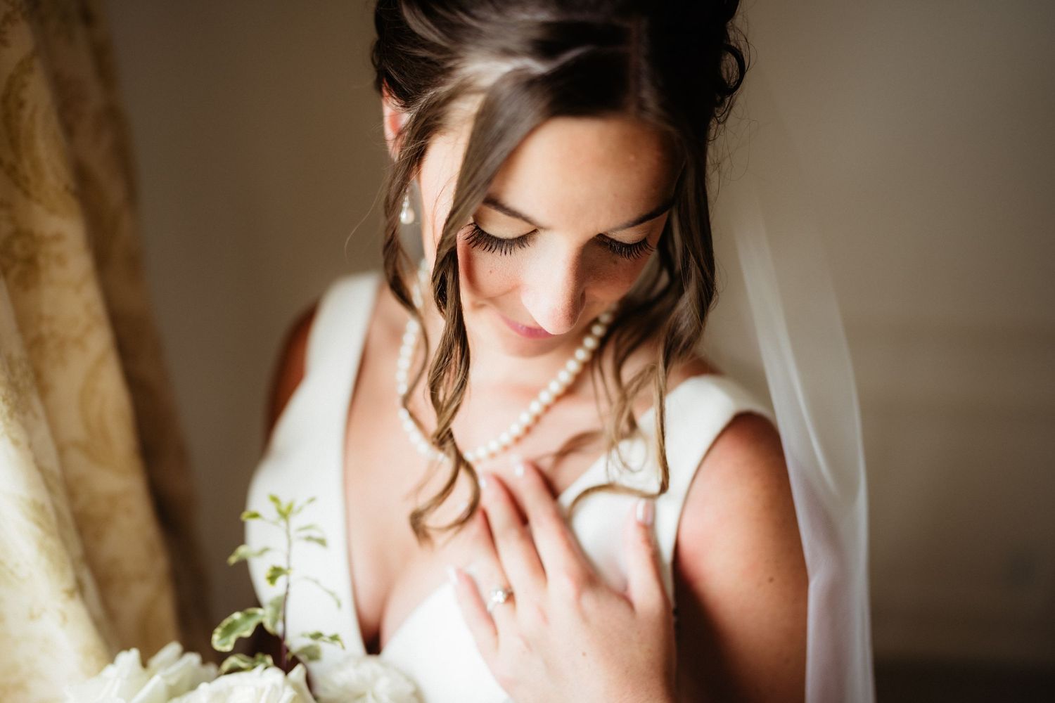 Bride in white gown holding white floral bouquet, wearing pearl necklace and ring, soft natural light.