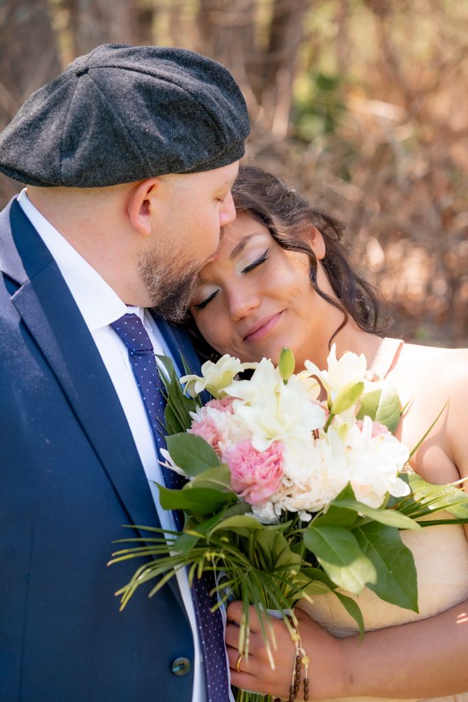 Bride and groom share a tender moment, holding a pink and white peony bouquet outdoors.