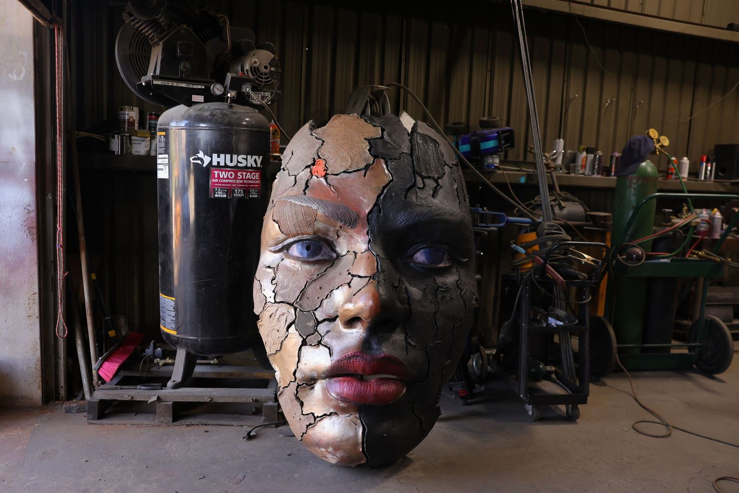 A cracked and damaged mask with dark features sits in a workshop with exercise equipment in the background.