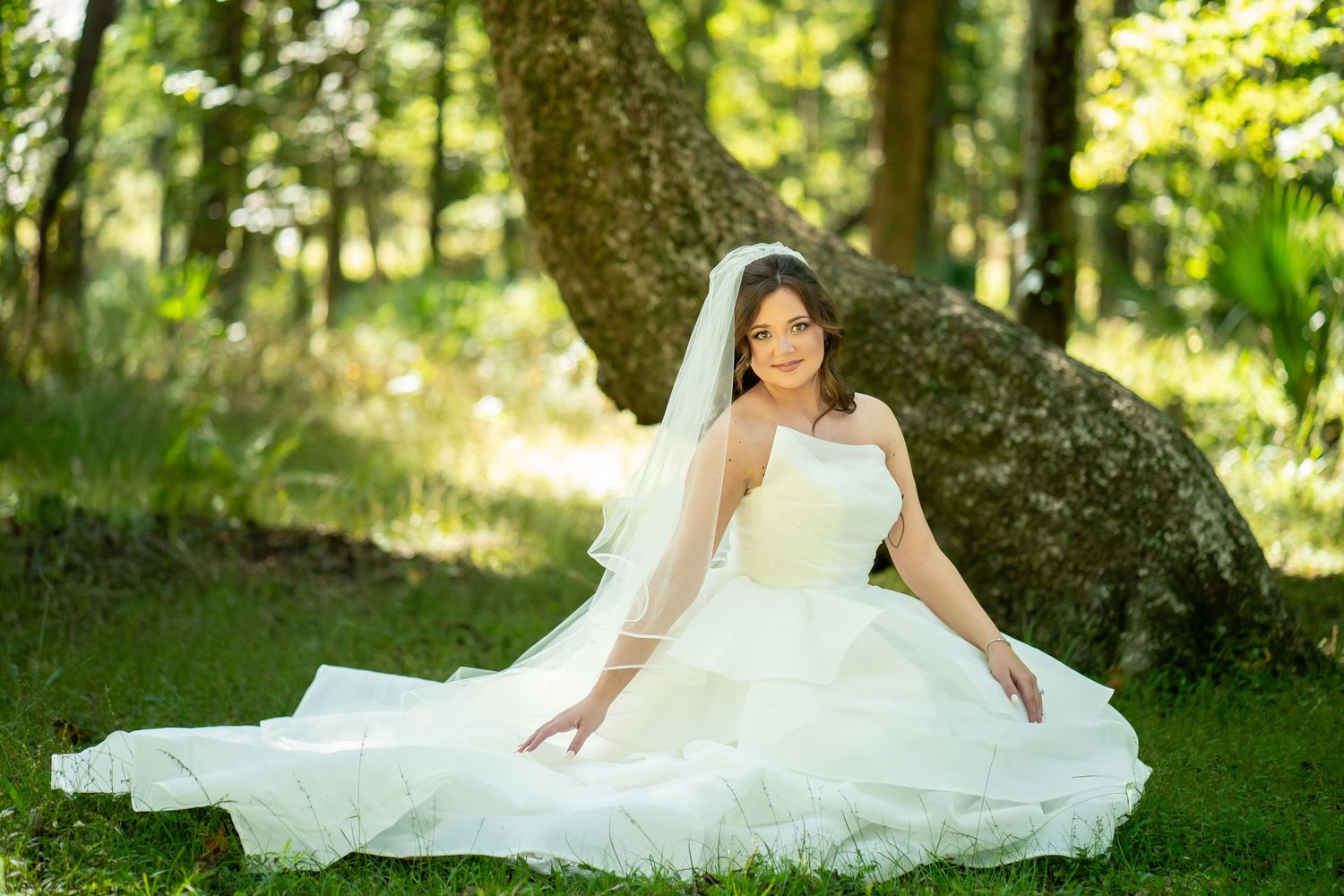 A bride in a flowing white gown reclines gracefully on green grass beneath towering trees in a sunlit forest setting.