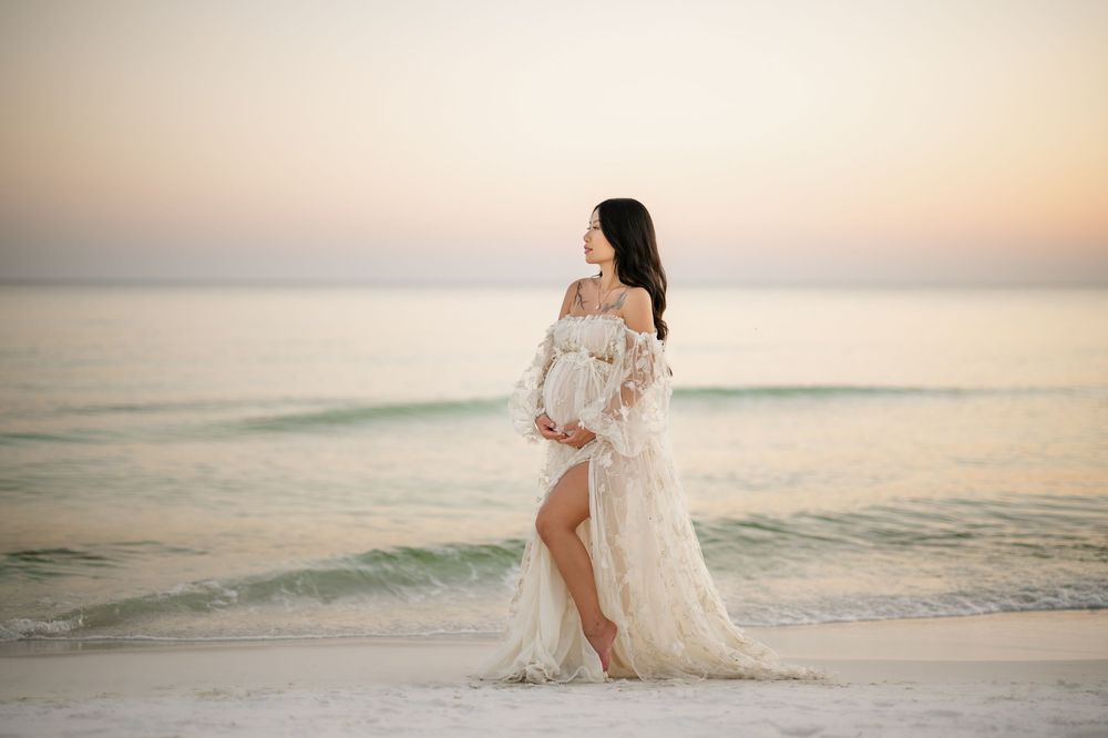 A woman in a flowing white lace dress stands barefoot on a beach at sunset with calm ocean waters in the background.