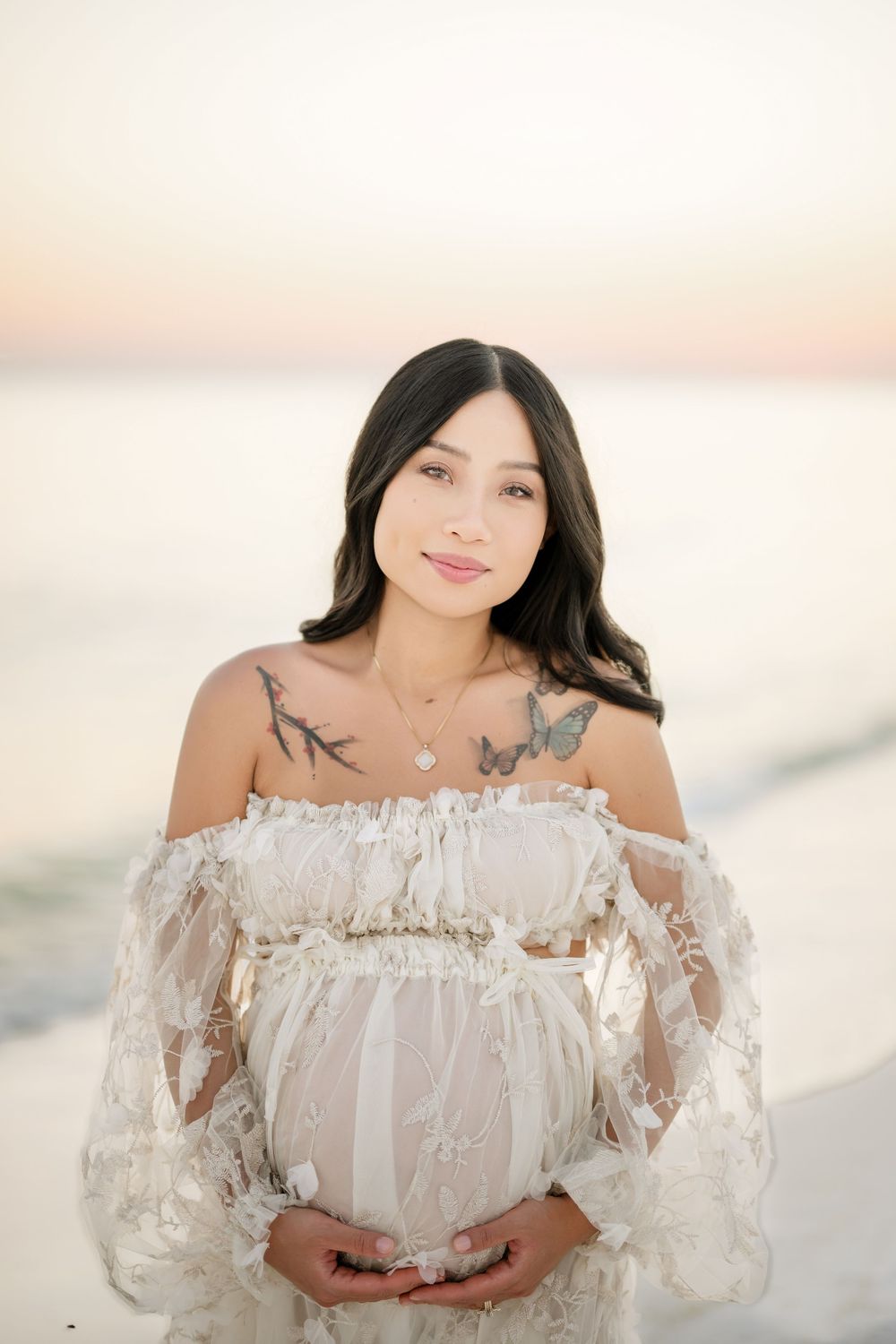 A person in a romantic off-shoulder white lace maternity dress poses outdoors at sunset with a soft glow.