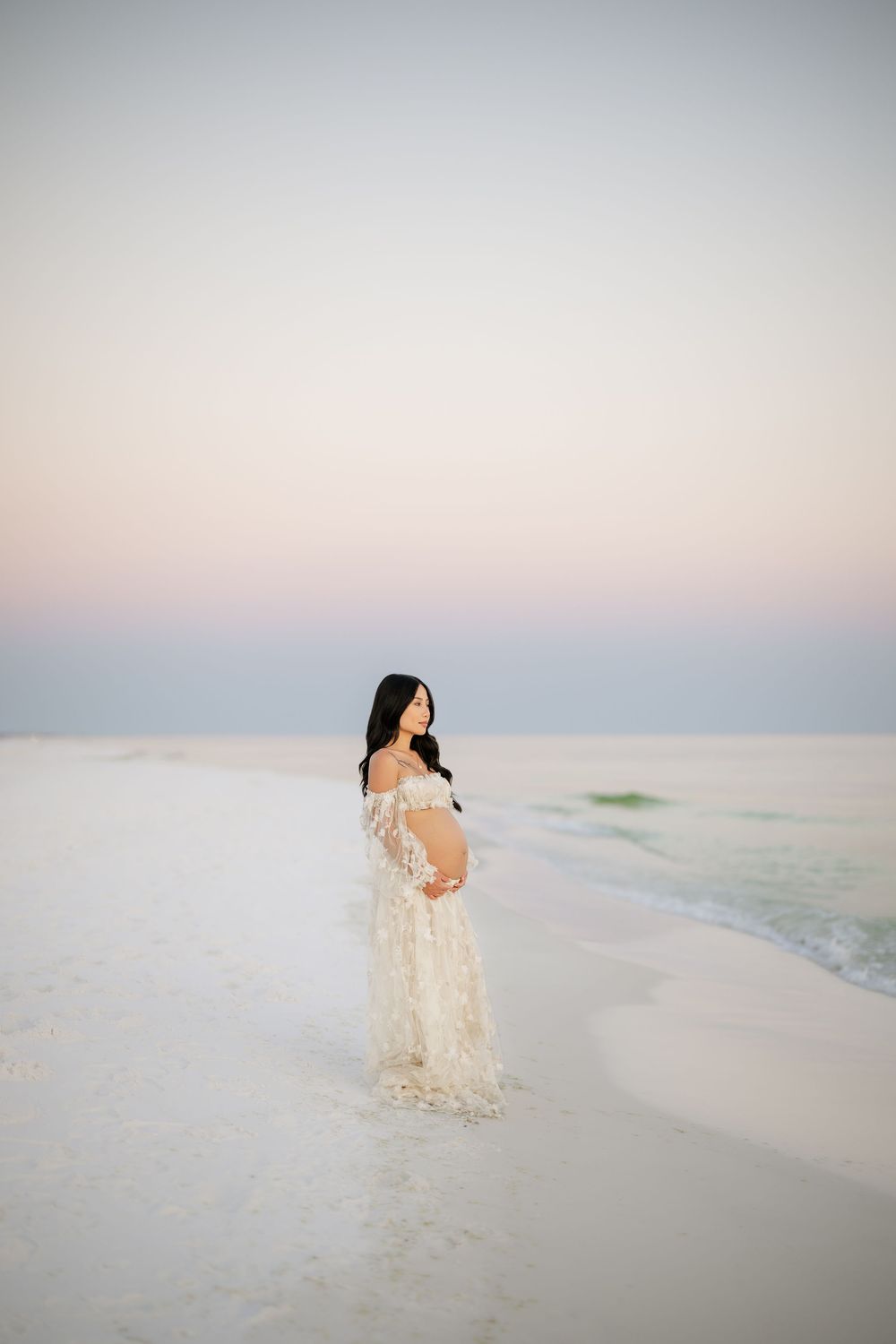 A person in a flowing cream lace dress poses gracefully on a serene white sand beach at sunsetPanama City beach maternity photoshoot, best