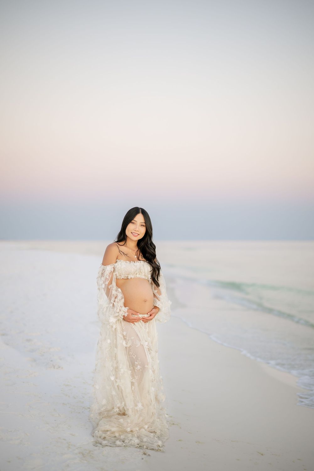 A figure in a flowing lace robe poses gracefully on a misty white sand beach at sunset.
